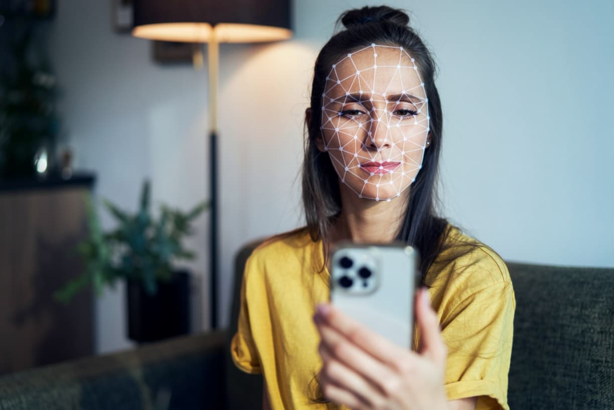 A woman holding her phone up to her face while a white landmark mesh maps her features, illustrating face verification with anti-spoofing.
